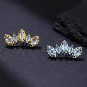 Può includere: Due paia di orecchini color oro e argento. Ogni orecchino presenta un gruppo di pietre preziose blu chiaro, taglio marquise. Gli orecchini sono esposti su una superficie blu scuro con un accento nero strutturato.