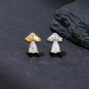 以下が含まれることがあります： キノコ型のスタッドピアス2つ。片方は金色のキャップと、茎にクリアな洋梨型の石が付いています。もう片方は銀色のキャップと、茎にクリアな洋梨型の石が付いています。暗い背景にあります。
