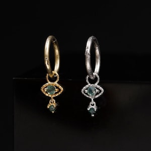 Op de afbeelding: Twee oorringen, één goudkleurig en één zilverkleurig, elk met een bungelende bedel in de vorm van een oog met een kleine, donkergroene edelsteen in het midden en een minuscuul bijpassend steentje onderaan het oogmotief.