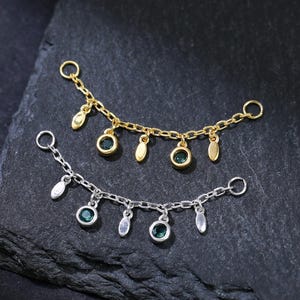 May include: Two delicate chain jewelry pieces, one gold-toned and one silver-toned, each adorned with small dangling charms and gemstones. The gold piece features green gemstones and teardrop charms, while the silver piece has blue-green gemstones and elongated charms.