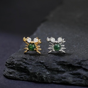 May include: A pair of crab-shaped stud earrings. One earring is gold-toned with white accents, the other is silver-toned. Both have a round, green gemstone center. The earrings are displayed on a dark, textured surface.