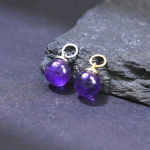 Può includere: Due piccoli orecchini rotondi in ametista viola intenso con anelli color oro. Gli orecchini sono esposti su una superficie strutturata grigio scuro su uno sfondo nero. Le pietre preziose hanno una superficie lucida e riflettente.