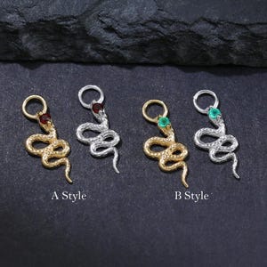 Op de afbeelding: Vier slangen-vormige bedels in goud- en zilverkleur, elk met een edelsteen accent. Twee bedels zijn gelabeld "A Style" en twee "B Style". De edelstenen zijn rood en groen. Op een donkere achtergrond.