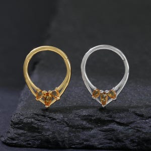 Puede incluir: Dos piercings nasales, uno dorado y otro plateado, se muestran sobre un fondo oscuro. Cada anillo presenta un diseño de lágrima con tres pequeñas piedras preciosas de color ámbar en la parte inferior. Los anillos se muestran en primer plano.