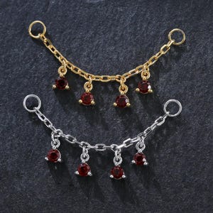 May include: Two delicate chain jewellery pieces. One is gold-toned, the other silver-toned. Each features small, dangling red gemstones. The chains are displayed on a dark, textured surface, showcasing the jewellery's design and colour contrast.