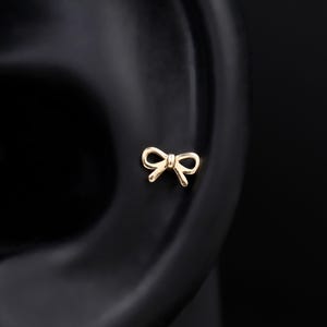 May include: A small, gold-toned stud earring shaped like a delicate bow. The earring features a polished finish and a simple, symmetrical design with two loops and two short, downward-pointing tails, positioned on a dark, neutral ear model.