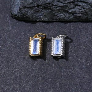 Peut inclure: Deux pendentifs rectangulaires avec des finitions dorées et argentées, chacun orné d'une pierre bleu clair. Les pendentifs sont présentés sur une surface gris foncé, avec une roche gris foncé en arrière-plan. Les pendentifs sont petits et délicats.