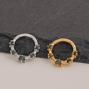 May include: Two circular nose rings. One is silver-toned, the other gold-toned. Both have small, dark green gemstones set around the outer edge. The jewellery is displayed on a dark grey surface.