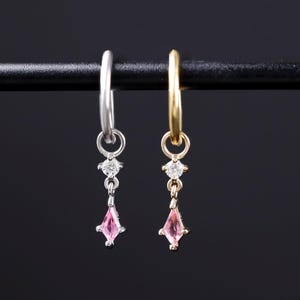May include: Two hoop earrings, one silver-toned and one gold-toned, each featuring a small clear round stone and a dangling pink kite-shaped stone. The earrings are suspended from a thin horizontal bar against a dark background.