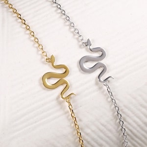 May include: Two delicate snake chain bracelets. One is gold-toned, the other is silver-toned. Each bracelet features a stylized snake pendant. The bracelets are displayed on a white, textured surface.