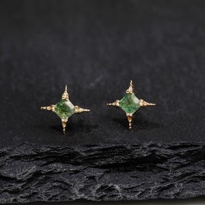 May include: A pair of gold stud earrings featuring a square-cut green gemstone at the center. Each earring is designed with four delicate, pointed metal prongs extending outward from the stone, creating a star-like shape with a metallic finish.