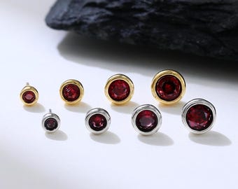May include: Assortment of small, round earrings with red gemstones and gold or silver settings. The earrings are arranged in a row on a white surface.