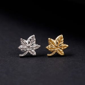 May include: Two stud earrings shaped like maple leaves, one finished in a bright silver tone and the other in a warm gold tone. Both earrings feature intricate, etched vein detailing across the surface of the leaf-shaped metal design.