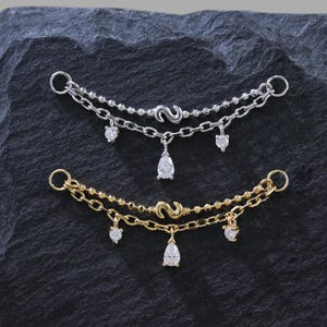 Puede incluir: Se muestran dos delicadas cadenas corporales, una plateada y otra dorada. Cada cadena presenta un diseño de doble hebra con pequeños acentos brillantes y un dije en forma de lágrima. Las cadenas se presentan sobre una superficie oscura y texturizada.