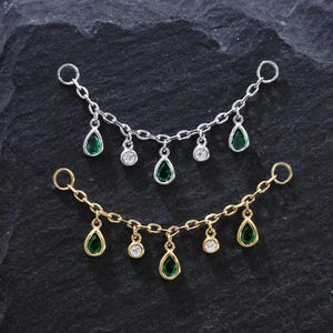 Puede incluir: Dos delicados collares de cadena. Uno es plateado, el otro dorado. Cada uno presenta gemas verdes en forma de lágrima y pequeñas gemas redondas y transparentes. Los collares se exhiben sobre una superficie oscura y texturizada.