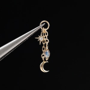 May include: A gold charm featuring a small star with a central clear stone, a delicate chain, a small oval iridescent stone, and a crescent moon pendant. The charm is held by silver-toned metal tweezers against a solid black background.