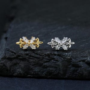 Puede incluir: Dos anillos con forma floral, uno dorado y otro plateado, cada uno adornado con piedras preciosas transparentes. Los anillos se exhiben sobre una superficie oscura y texturizada, creando un contraste que resalta su intrincado diseño.