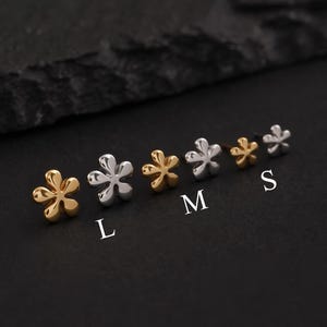 May include: A collection of flower-shaped stud earrings in gold and silver tones. The earrings are arranged in a row, with sizes labeled L, M, and S. The background is a dark, textured surface.