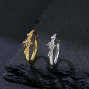 May include: Two delicate rings, one gold and one silver, each featuring a star-shaped design with a central gemstone. The rings are displayed on a dark, textured surface, creating a contrast that highlights their intricate details. Jewelry.