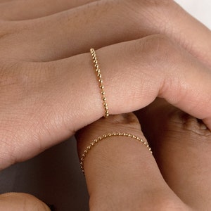 Puede incluir: Dos anillos de oro con cuentas en los dedos de una persona. Los anillos son finos y delicados, con pequeñas cuentas espaciadas uniformemente.