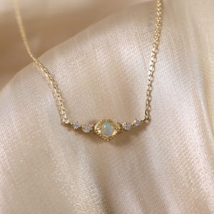 Op de afbeelding: Een delicate gouden ketting met een centraal, vierkant gevormde opaal hanger, geflankeerd door kleinere, ronde edelstenen. De ketting is geplaatst tegen een achtergrond van zachte, gouden stof, wat het elegante ontwerp benadrukt.