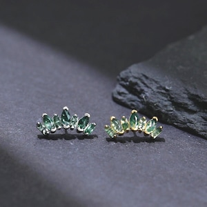 May include: Two pairs of earrings, each featuring a row of green, teardrop-shaped gemstones. One pair has a silver-colored setting, while the other has a gold-colored setting. The earrings are displayed on a dark gray surface.