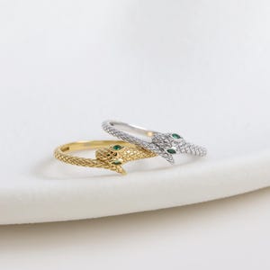 May include: Two snake rings, one gold and one silver, rest on a white surface. The gold ring features a textured band and green gemstone eyes. The silver ring is encrusted with small, clear stones and has green gemstone eyes.