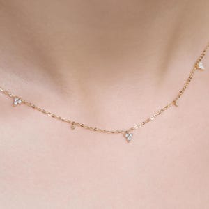 May include: A delicate gold necklace featuring small, sparkling diamond accents. The necklace has a fine chain and several tiny diamond pendants, with a larger cluster of diamonds near the center. The necklace is shown against a skin-toned background.