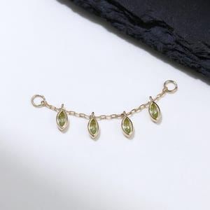 Puede incluir: Una delicada cadena de oro con cinco colgantes de piedras preciosas de color verde oliva. Los colgantes en forma de lágrima están espaciados uniformemente a lo largo de la cadena curva. La joya se encuentra sobre un fondo blanco con una superficie oscura y texturizada en la esquina superior derecha.