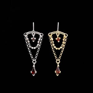 May include: Two ear jackets featuring a triangular chain design. One is silver-toned and the other is gold-toned. Each piece includes a small dark red gemstone at the top and a larger marquise-cut dark red gemstone at the bottom point.