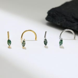 Puede incluir: Tres anillos de nariz con acentos de piedras preciosas verdes. Los anillos son de oro, plata y una barra recta.