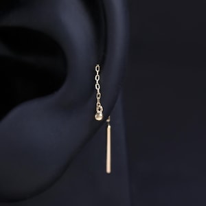 May include: A close-up of a gold earring on an ear. The earring features a delicate chain with a small ball at the end, and a straight bar. The earring is gold in color.