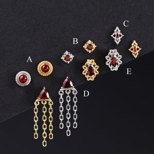 Puede incluir: Una colección de pendientes de botón y colgantes con piedras de color rojo intenso engastadas en metal dorado y plateado. Los diseños incluyen formas circulares, de estrella, ovaladas ornamentadas y triangulares con cadenas, etiquetadas de la A a la E.