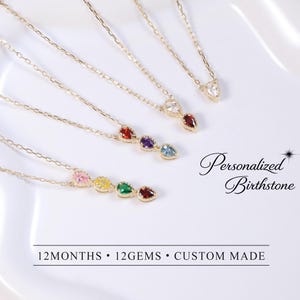 May include: Gold-toned necklaces with colorful gemstone pendants. The necklaces feature a delicate chain and a variety of birthstone colors. The image also includes the text "Personalized Birthstone" and "12 MONTHS • 12 GEMS • CUSTOM MADE".
