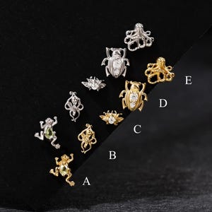 Puede incluir: Una colección de pequeños pendientes de botón de metal en tonos plateados y dorados, con formas de animales e insectos como ranas, pulpos, escarabajos y abejas. Algunas piezas tienen piedras claras o verde claro. Etiquetadas como A, B, C, D y E.