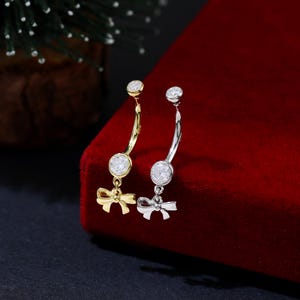 May include: Two belly button rings, one gold-toned and one silver-toned, each featuring a clear, round gemstone and a small bow charm. The curved bars are displayed against a red velvet surface, with a small Christmas tree in the background.