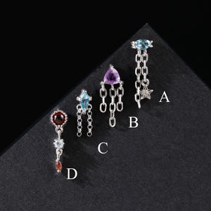 May include: Four silver-toned metal body jewelry pieces labeled A, B, C, and D. Piece A has a blue stone and star charm. Piece B features a triangular purple stone with hanging chains. Piece C has a blue kite-shaped stone. Piece D has a red stone.