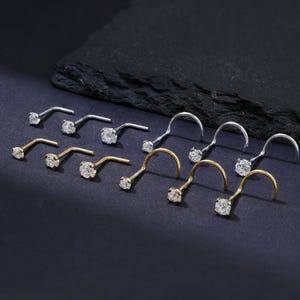 May include: An assortment of nose studs in gold and silver tones, each featuring a small, clear gemstone. The studs are arranged on a dark surface, showcasing their delicate design and sparkle. The curved posts are visible.