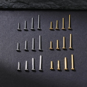 May include: A collection of small, stud earrings in silver and gold tones, arranged in rows on a dark surface. The earrings have a flat, circular front and a straight post. The studs vary in size.