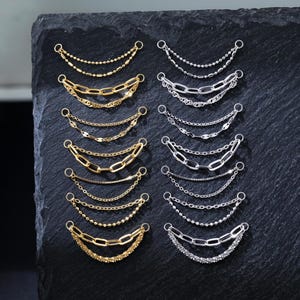 Puede incluir: Una variedad de colgantes de cadena dorados y plateados. Los colgantes presentan varios estilos de cadena, incluyendo cadenas de cuentas, de eslabones y de tipo curb. Cada colgante tiene un pequeño lazo en la parte superior para la fijación. Las cadenas se muestran sobre una superficie oscura y texturizada.