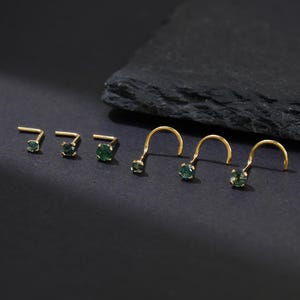 May include: A collection of gold nose studs and nose rings with round, green gemstones. The jewelry is displayed on a dark surface, showcasing the different styles and sizes available. The studs have a straight post, while the rings have a curved design.