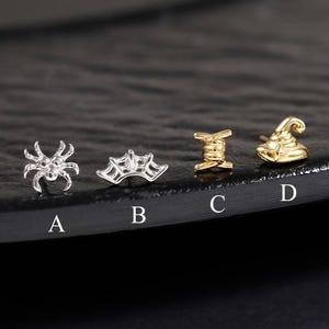Op de afbeelding: Vier kleine oorknopjes gelabeld van A tot D. A is een zilveren spin, B is een zilveren spinnenweb, C is een goudkleurige klos garen en D is een goudkleurige puntmuts. Elk sieraad is uniek in vorm en kleur en staat in een horizontale rij.
