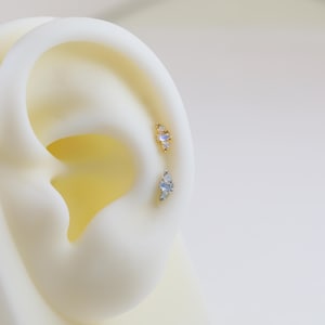 May include: Close-up of two ear piercings. The top earring is gold-toned with small, light blue stones. The bottom earring is silver-toned with similar light blue stones. Both earrings are set on a white ear model.
