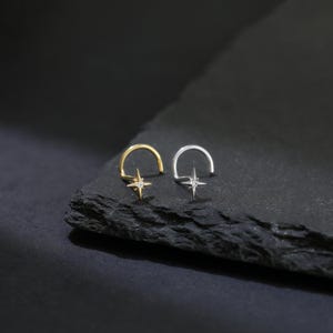 May include: Two nose rings, one gold and one silver, each featuring a starburst design with a central gemstone. The rings are displayed on a dark, textured surface, creating a contrast that highlights the jewelry's details.