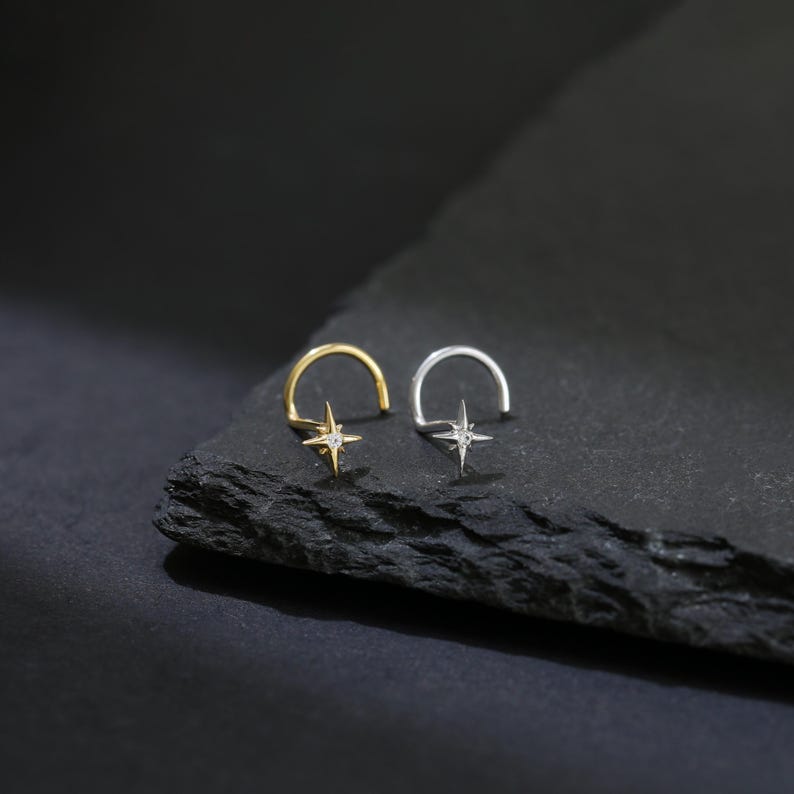 May include: Two nose rings, one gold and one silver, each featuring a starburst design with a central gemstone. The rings are displayed on a dark, textured surface, creating a contrast that highlights the jewelry's details.