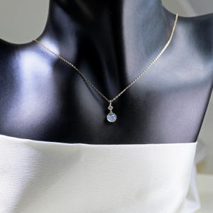May include: A delicate gold chain necklace with a pendant featuring a round, light blue gemstone and a smaller, square-shaped gemstone.