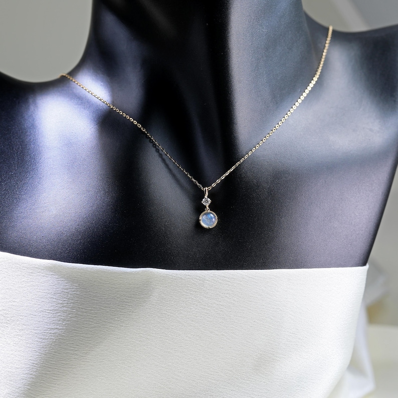 May include: A delicate gold chain necklace with a pendant featuring a round, light blue gemstone and a smaller, square-shaped gemstone.