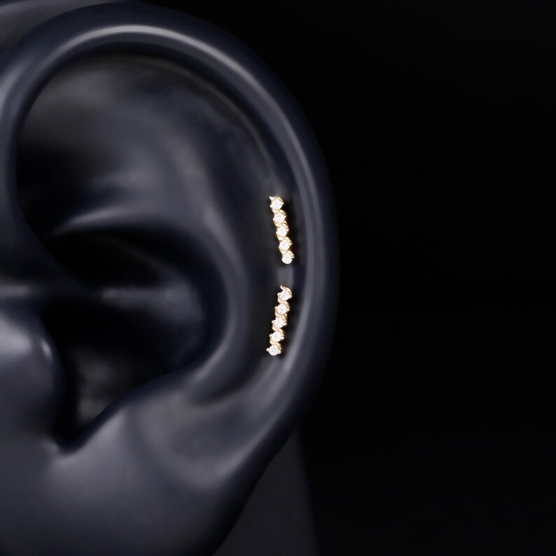 K&ouml;nnte beinhalten: Zwei goldene Ohrpiercings mit vertikalem Stabdesign, jeweils besetzt mit f&uuml;nf kleinen, klaren, rund geschliffenen Steinen in einer geraden Linie. Der Schmuck ist entlang des oberen Knorpelbereichs eines dunklen Ohrmodells platziert.