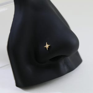 May include: A gold-colored nose stud in the shape of a four-pointed star with a small, clear gem in the center. The stud is set on a black nose form, showcasing the jewelry's design and detail. The starburst design adds a touch of elegance.
