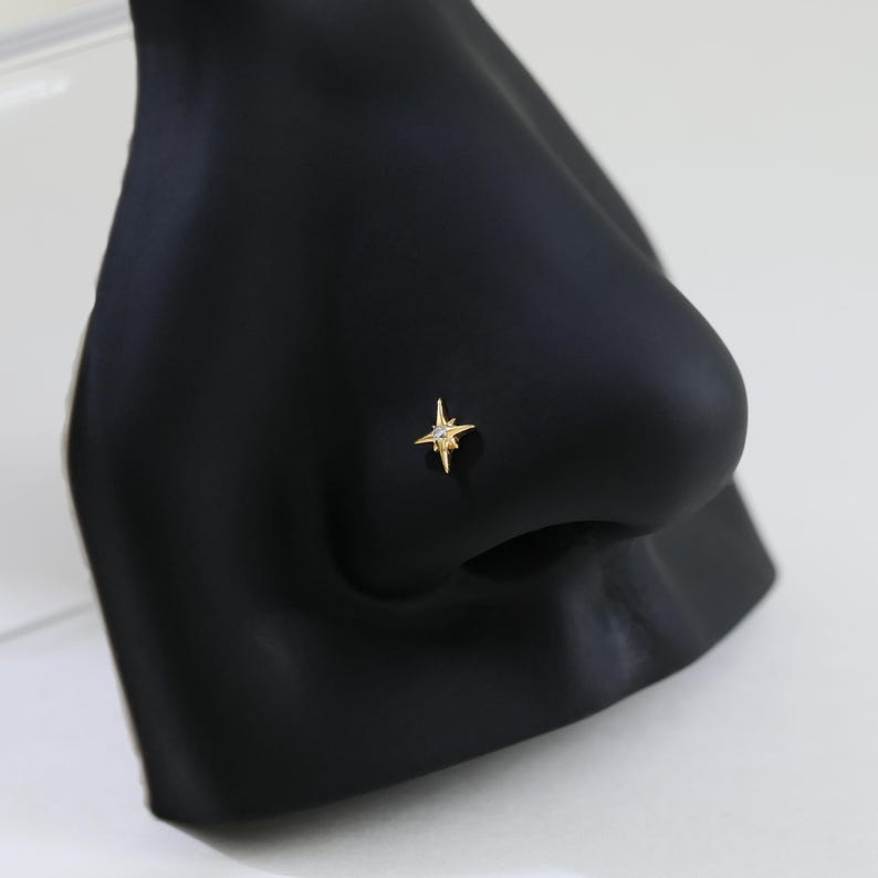 May include: A gold-colored nose stud in the shape of a four-pointed star with a small, clear gem in the center. The stud is set on a black nose form, showcasing the jewelry's design and detail. The starburst design adds a touch of elegance.